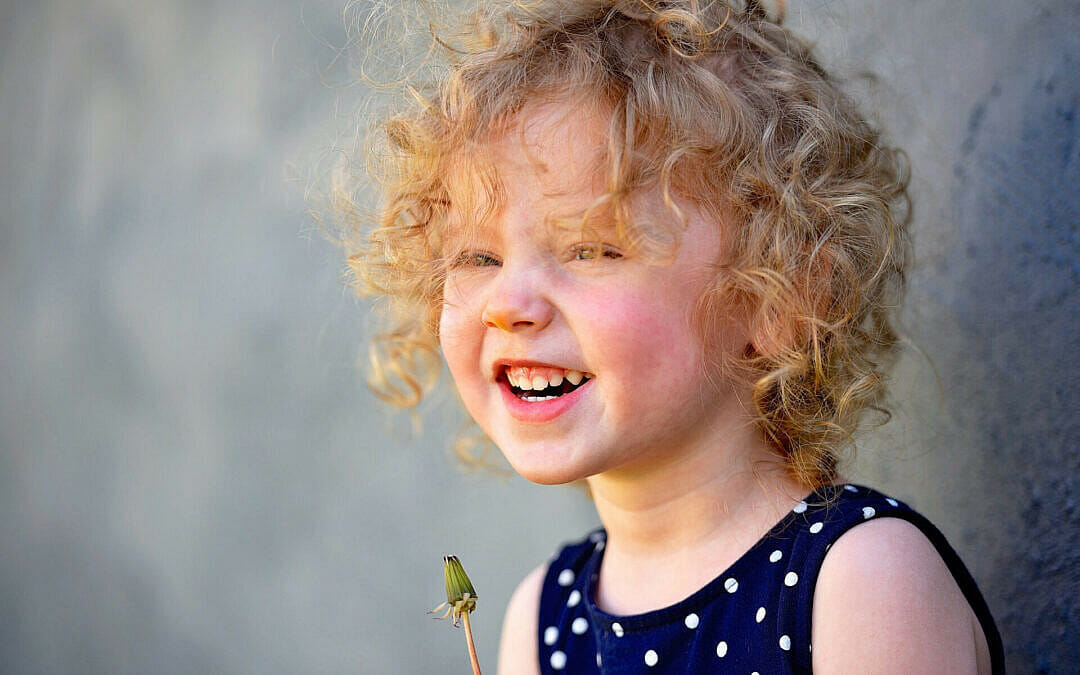 Smiling young child with curly blonde hair holding a dandelion during an outdoor portrait session